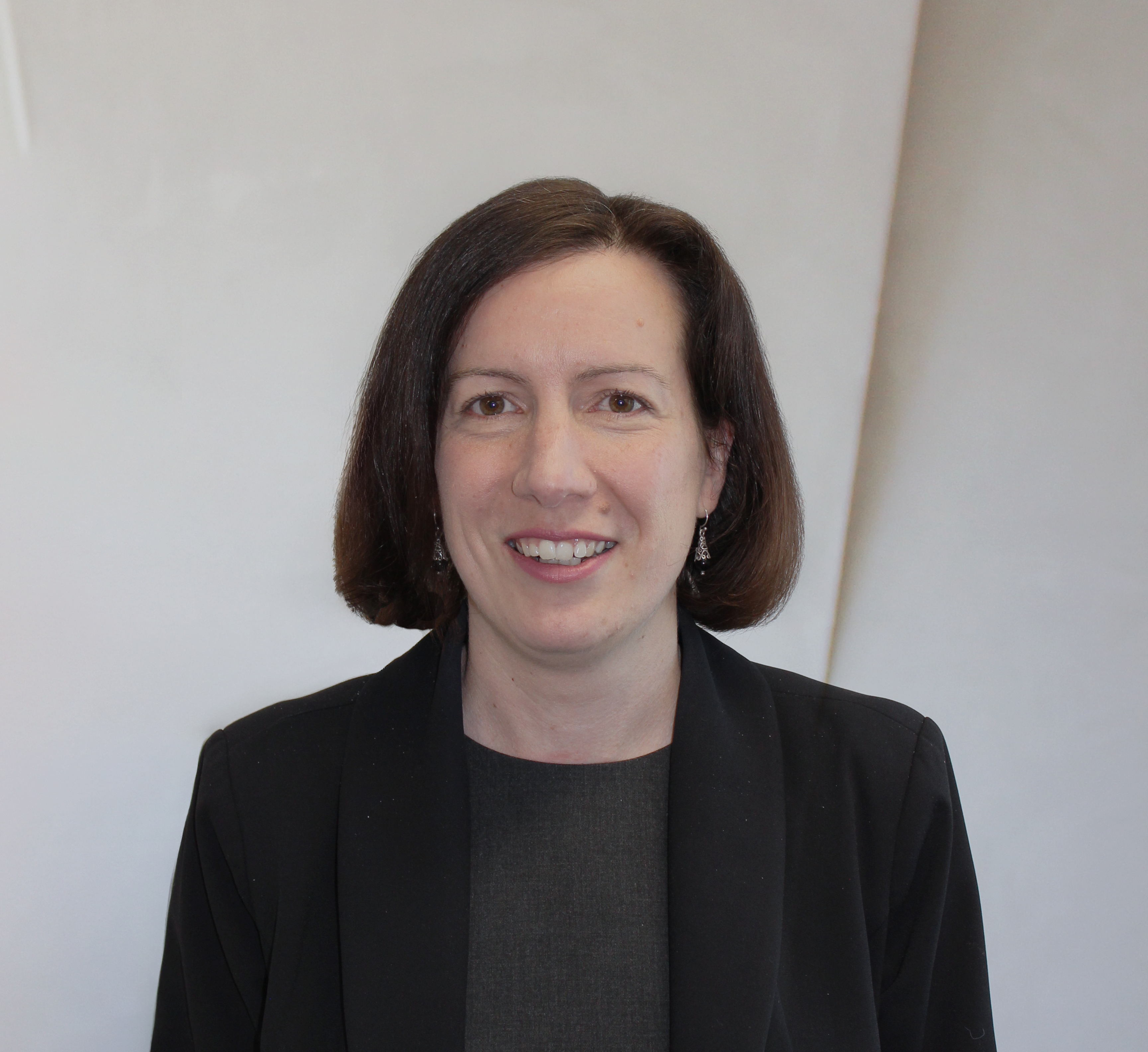 A woman with short, dark hair, wearing a black jacket with a grey shirt underneath, standing against a white background.