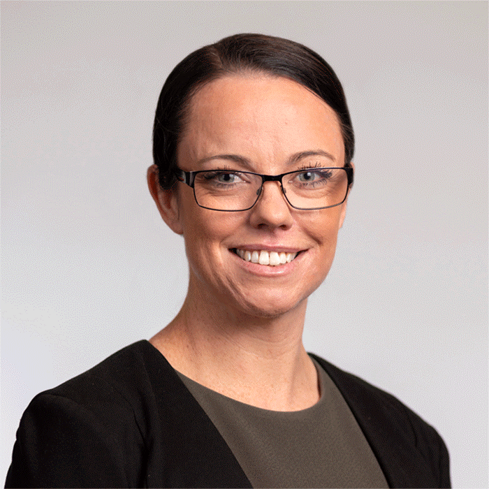 A woman with dark hair wearing glasses smiles at the camera. She is wearing dark clothing.