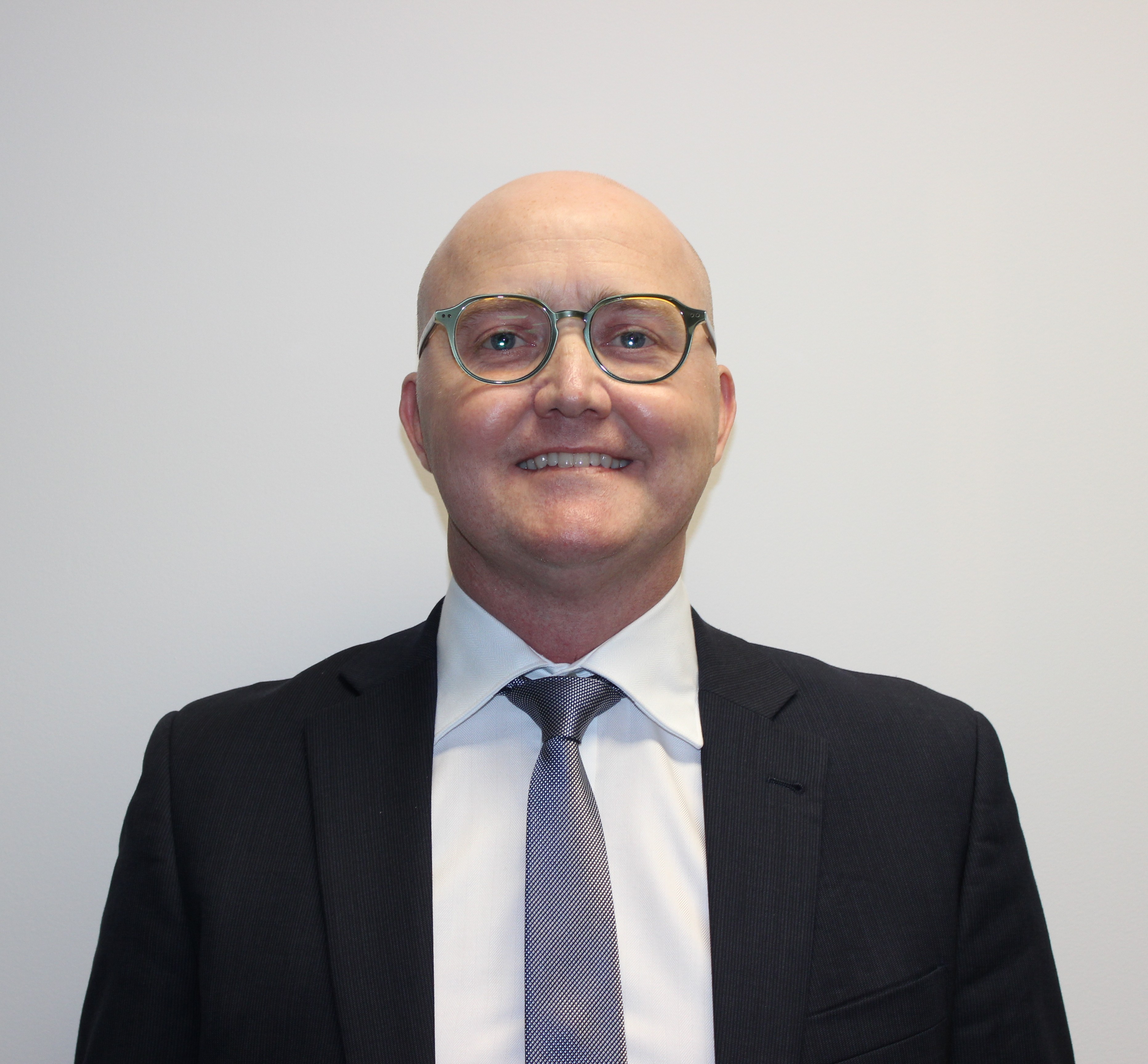 Portrait of a person wearing glasses, a dark suit jacket, white shirt and tie, facing the camera against a plain light background.