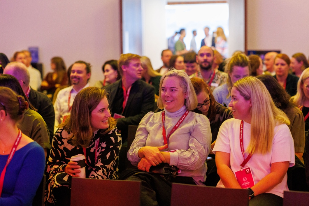A group of people are seated in a conference set-up talking amongst themselves while they wait for presentations to begin.