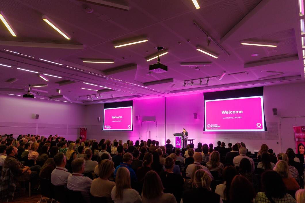 A speaker stands at a podium in a large, modern conference room filled with attendees. Two screens display a pink ‘Welcome’ slide.