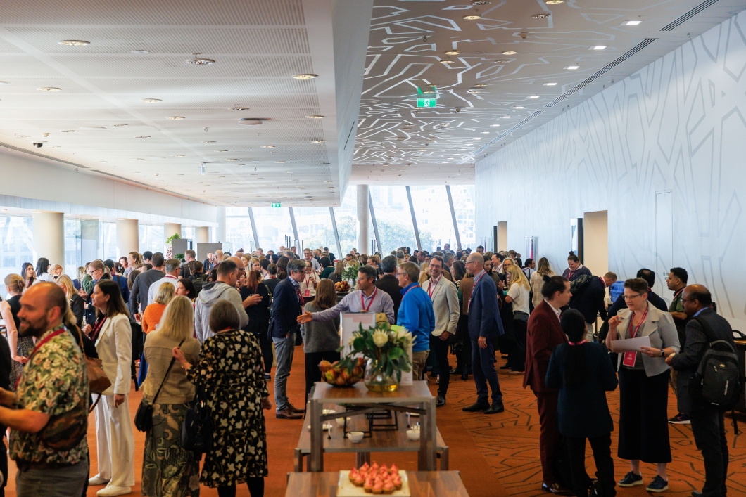 A crowded conference hallway with attendees networking, enjoying refreshments, and engaged in discussions, surrounded by modern architecture.
