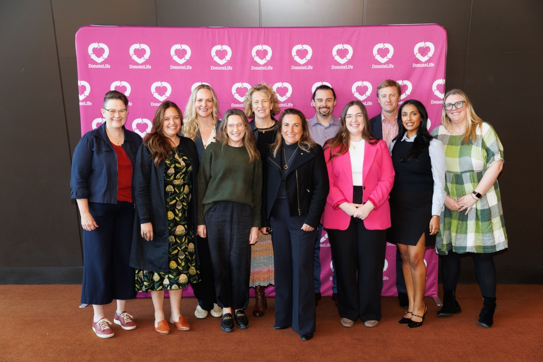 A group of 11 people pose and smile for a photo in front of a pink banner with the DonateLife logo.