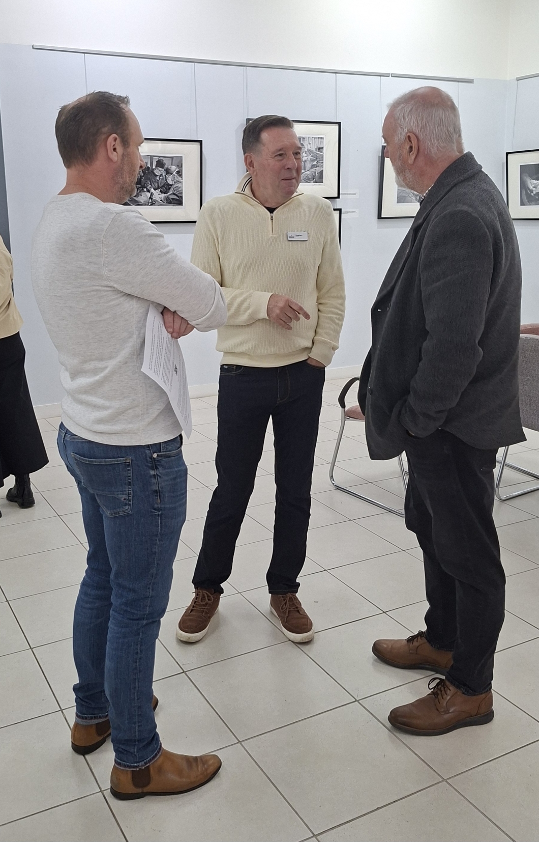 3 people stand and converse in an art gallery with framed black-and-white photographs on the walls. One person holds a piece of paper, and all are dressed casually.