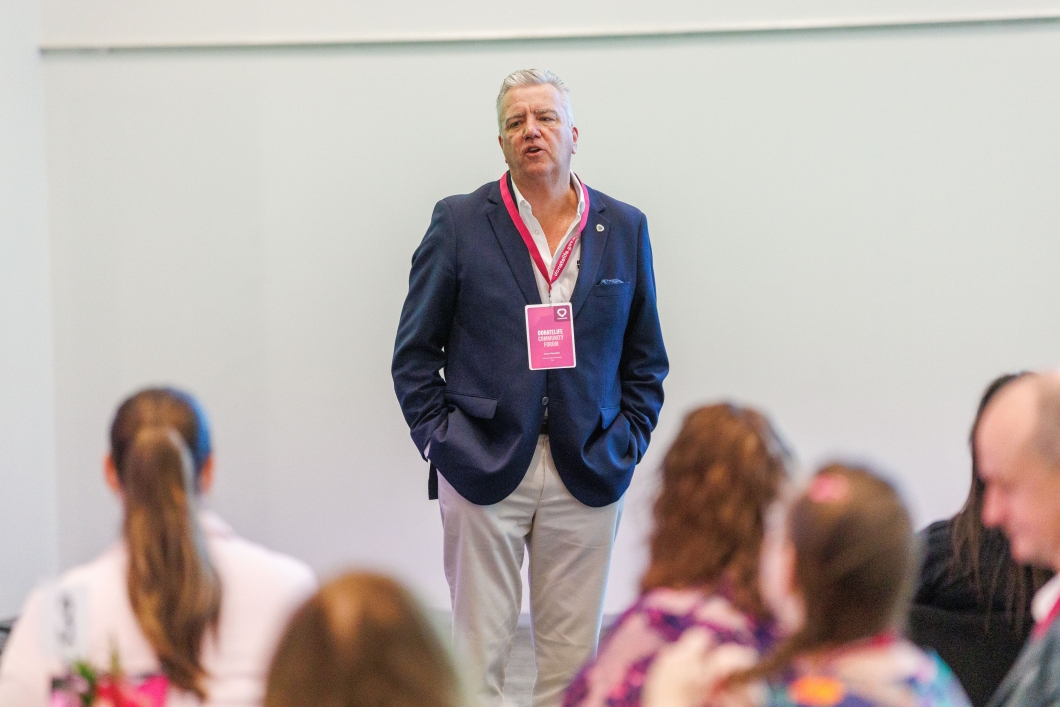 A man in a blue jacket stands in front of seated attendees, mid-sentence as he speaks to them.