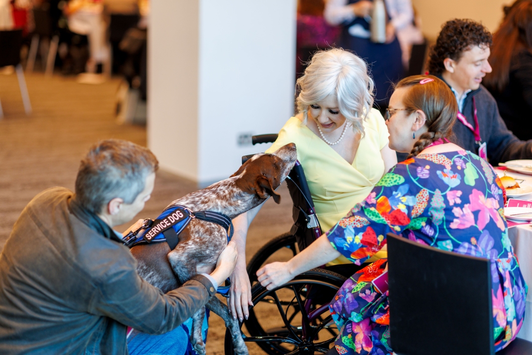 A service dog interacts with a woman in a wheelchair while seated at an event, with a woman in a brightly coloured dress sitting next to her, and a man kneeling on the floor petting the dog.