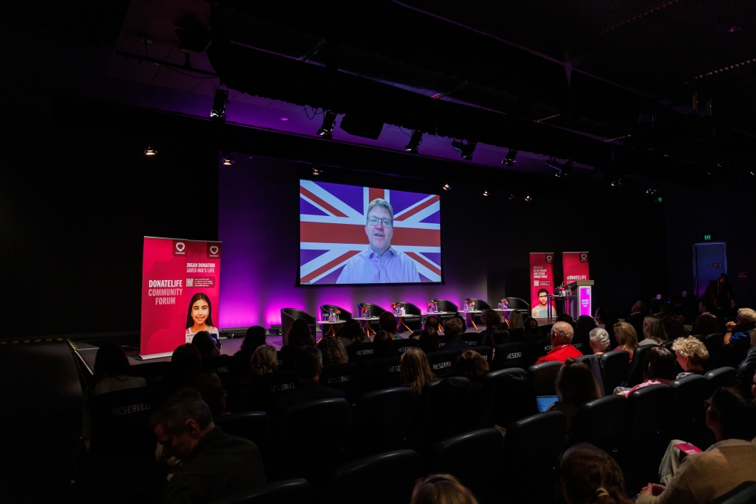  A conference setting with an audience seated, a large screen displaying a blurred speaker with a British flag, and informative banners.