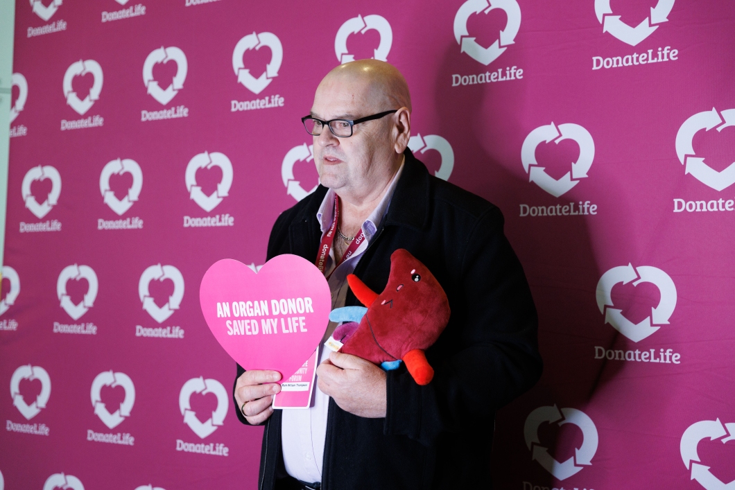 A person holds a heart-shaped sign reading ‘An organ donor saved my life’ and a plush liver against a pink DonateLife backdrop.
