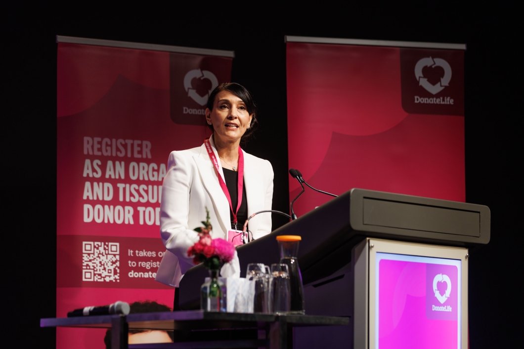 A speaker at a podium is mid-speech, surrounded by DonateLife banners and a floral arrangement on the table.
