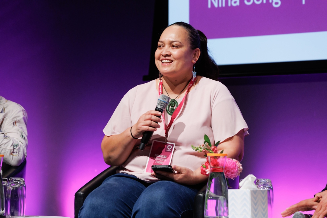 A woman in a light pink blouse holds a microphone while seated on stage, with a floral arrangement and a water pitcher nearby. 