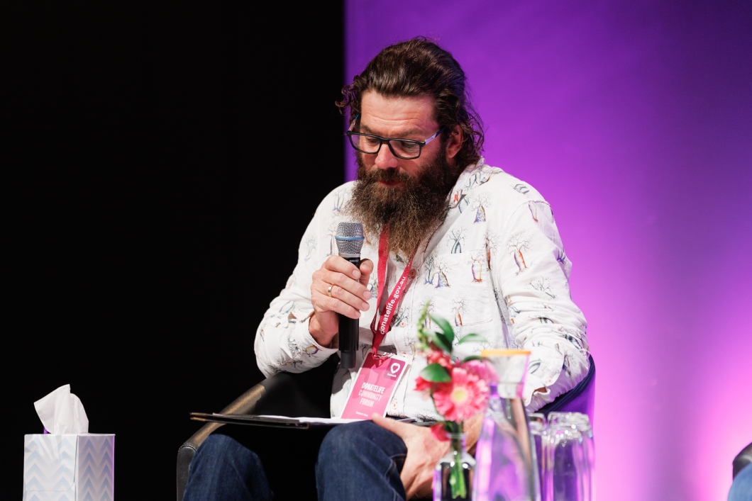 A speaker with a microphone dressed in a patterned shirt sits on stage, flanked by a glass of water and flowers, during a conference.