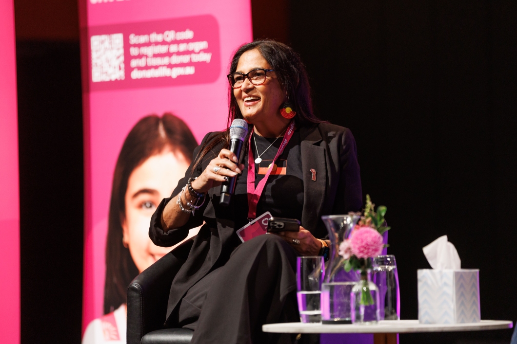 A speaker holds a microphone while seated in front of a pink backdrop, surrounded by a vase of flowers and a water jug on a table.