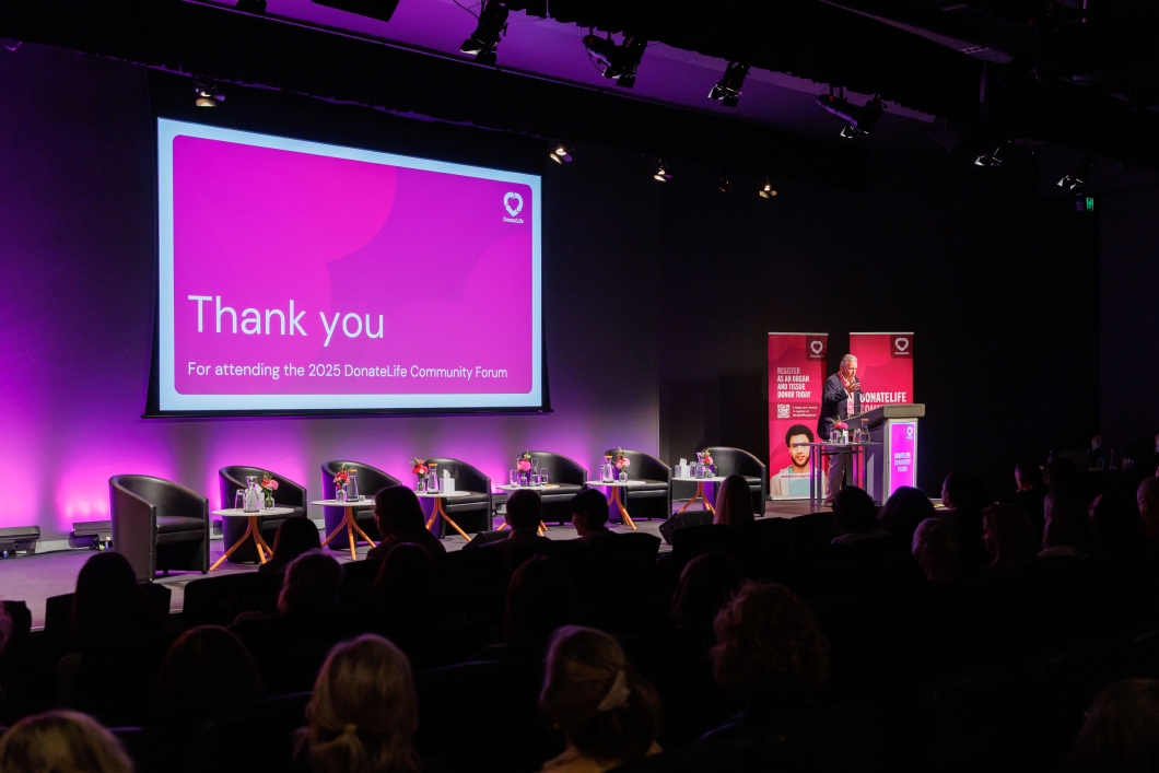 A speaker stands at a podium in a theatre style room filled with attendees. A screen displays a pink ‘Thank you’ slide.