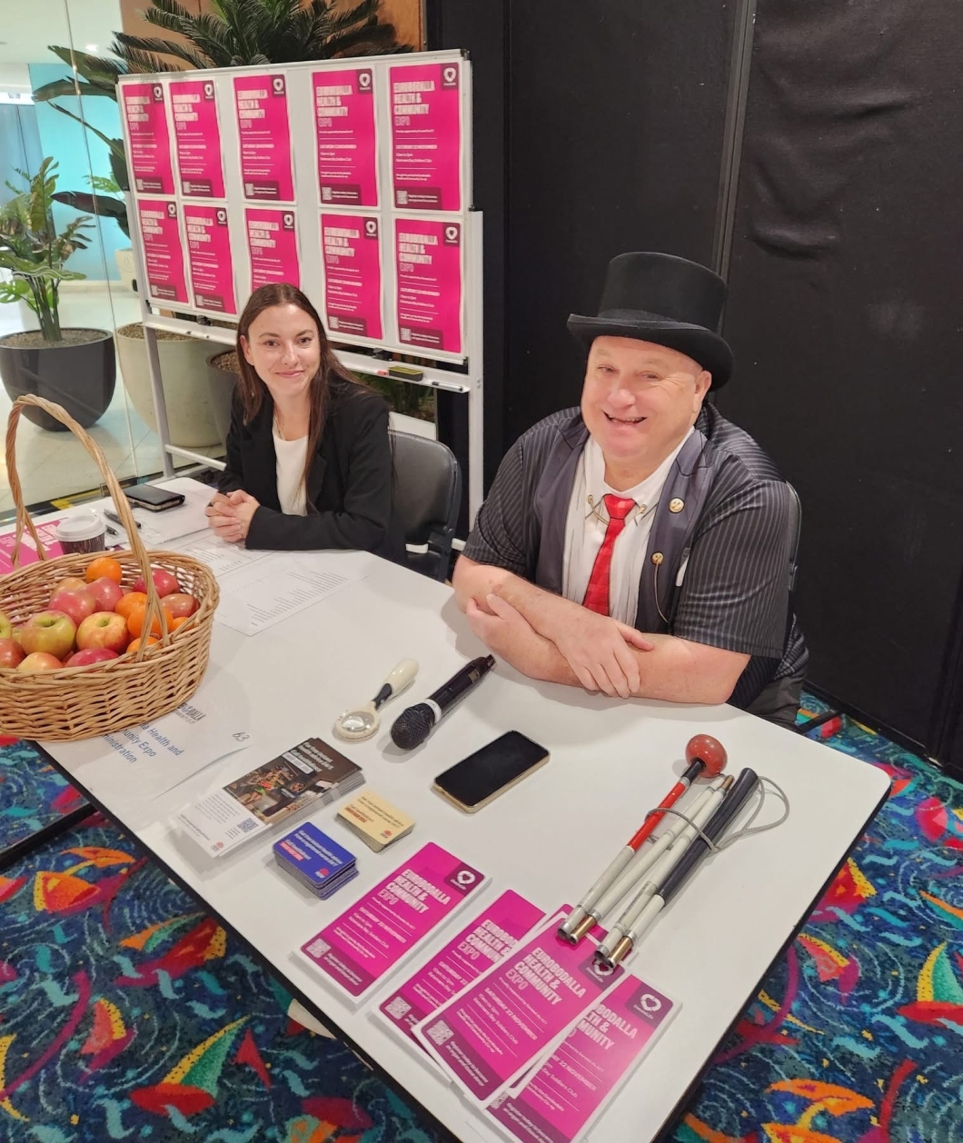 2 people sit behind an information table at an event. The table is covered with promotional materials, brochures, business cards, a phone, and a mobility cane. A woven basket filled with apples sits on one side of the table. Behind them is a display wall filled with bright pink posters. The setting features colourful patterned carpet and indoor plants.