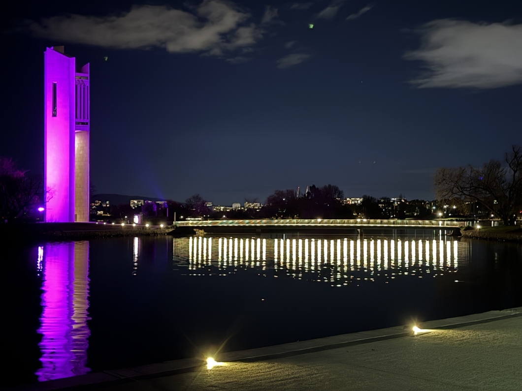 The National Carillon on Lake Burley Griffin is illuminated in bright magenta at night. The illuminated tower is reflected in the calm water, while lights from a nearby bridge and the Canberra skyline create reflections across the lake under a dark, partly clouded sky.