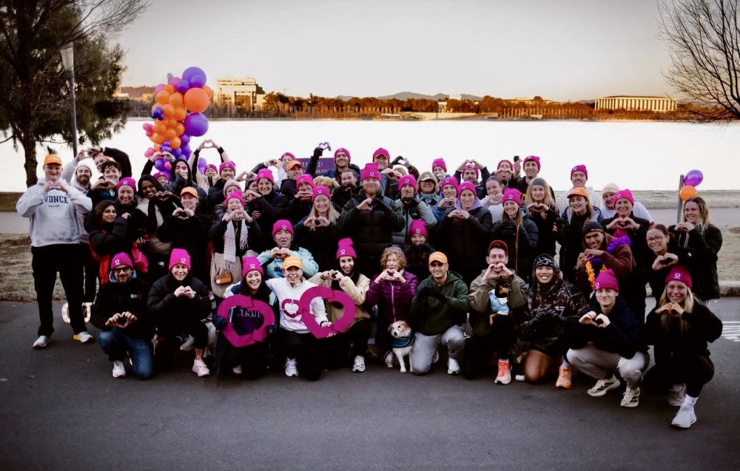 A large group of DonateLife supporters gathers by Lake Burley Griffin at sunrise for a community awareness walk. Participants wear bright magenta DonateLife beanies and form heart shapes with their hands for a group photo. Purple and orange balloons, DonateLife signs and a small dog are visible in the front row, with Canberra landmarks and the lake in the background.