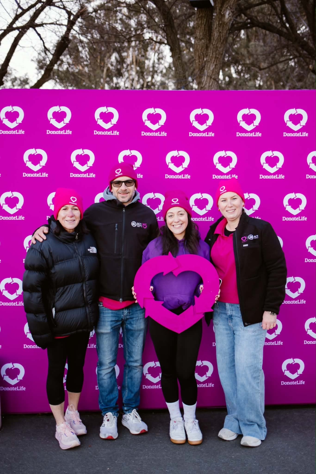 Four DonateLife supporters wearing bright magenta DonateLife beanies pose in front of a DonateLife media wall printed with the DonateLife logo. One person in the centre holds a large DonateLife heart symbol prop.&nbsp;
