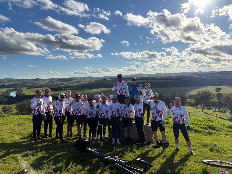 A group of cyclists wearing matching DonateLife Cycling Challenge jerseys poses for a photo on a hilltop overlooking rolling countryside. Several bicycles are resting on the grass in the foreground, while expansive green fields, a winding river and a sky filled with scattered clouds create a scenic backdrop. The cyclists are gathered around a survey marker at a rest stop during the challenge.