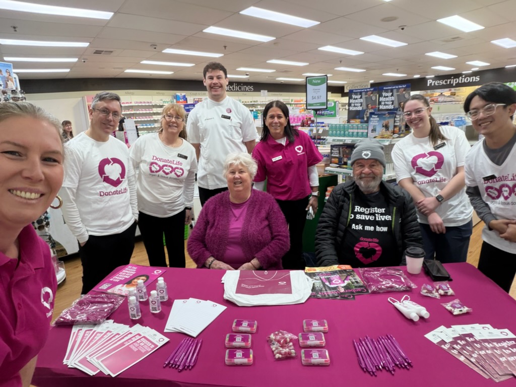  DonateLife volunteers and pharmacy staff stand behind a DonateLife information table inside a pharmacy. The table is covered with magenta promotional materials, including brochures, pens, badges and registration information. Several volunteers wear DonateLife shirts featuring the DonateLife logo and messages about organ and tissue donation, while pharmacy shelves and prescription signage are visible in the background. 
