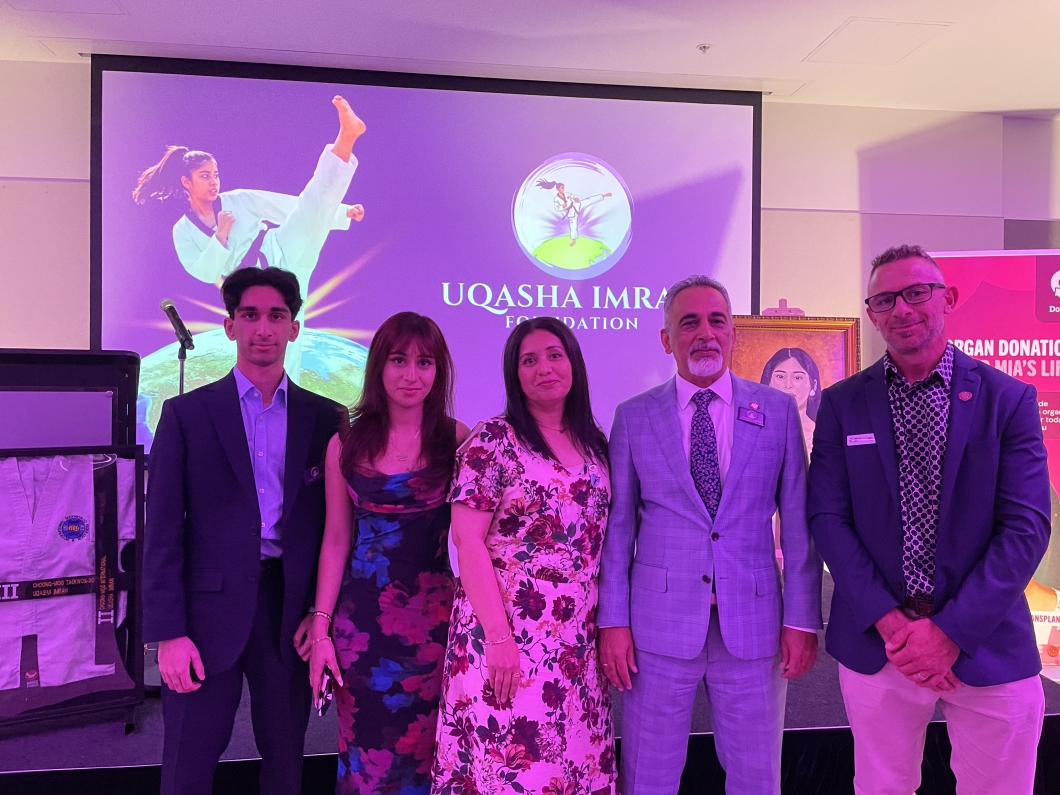 5 people standing side by side at an indoor event stage under pink and purple lighting, in front of a large screen displaying ’UQASHA IMRAN FOUNDATION’ and an image of a martial artist performing a high kick. A microphone stand and framed display are visible to the left, and a sign about organ donation appears on the right.