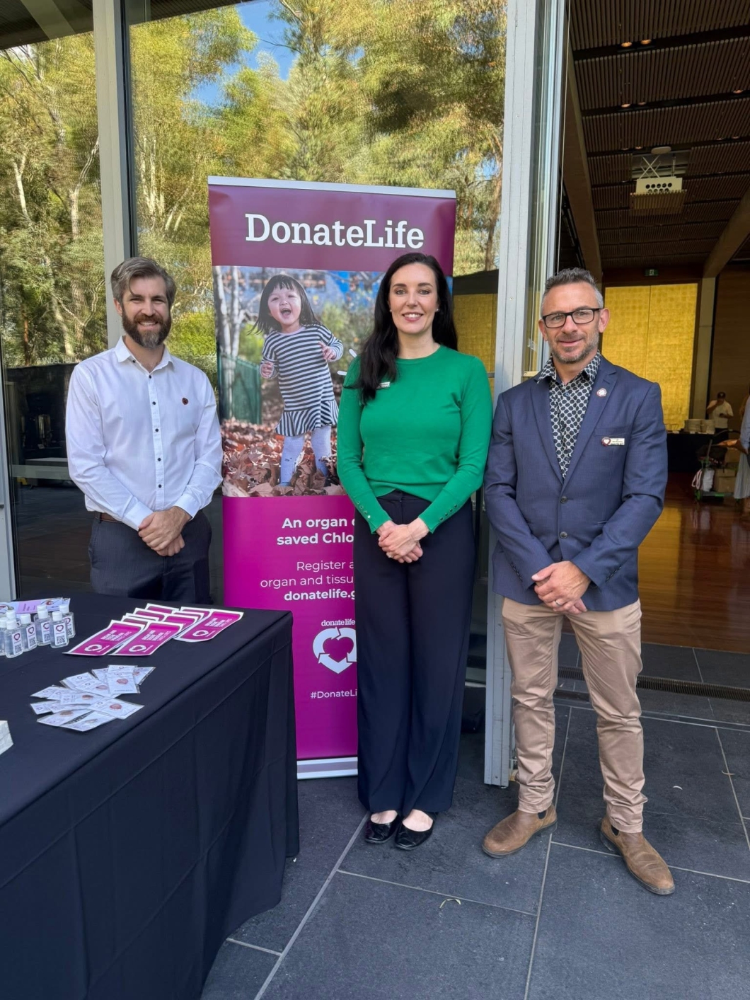 3 people stand in front of a DonateLife display banner near an open glass doorway. The banner shows a child walking through autumn leaves and includes DonateLife branding and registration messaging. A table covered with a black cloth is set up beside them, displaying DonateLife brochures and promotional items. Trees and greenery are visible in the background outside.