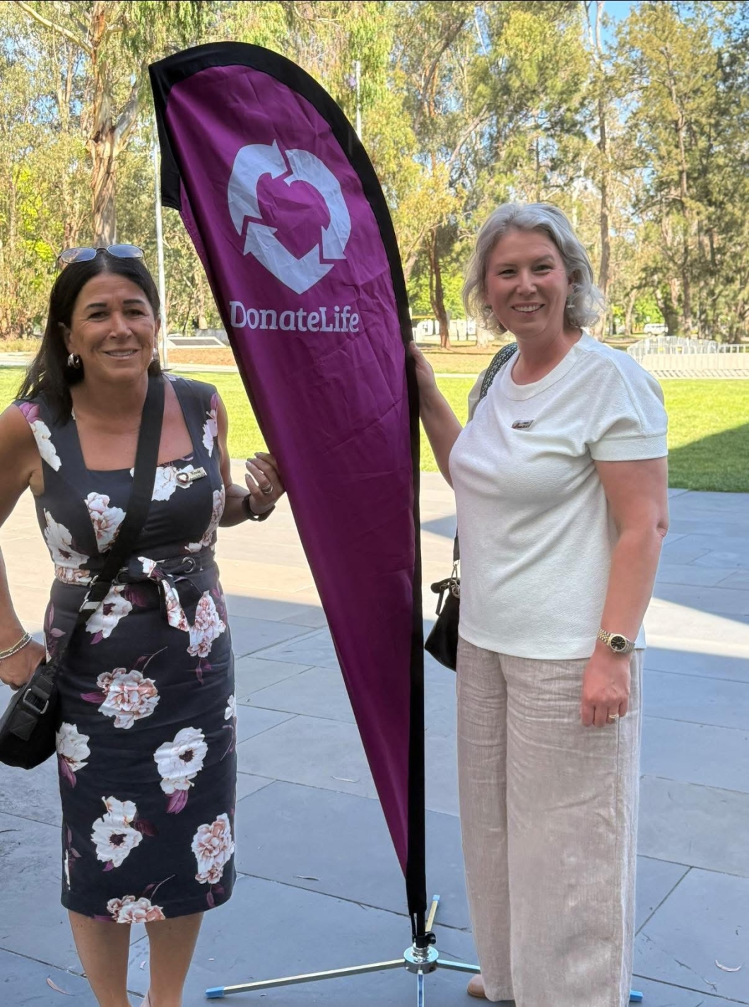 2 people stand outdoors beside a tall magenta DonateLife banner. The banner features the DonateLife logo and is set up on a metal base. The pair stand on a paved area with trees and greenery visible in the background.