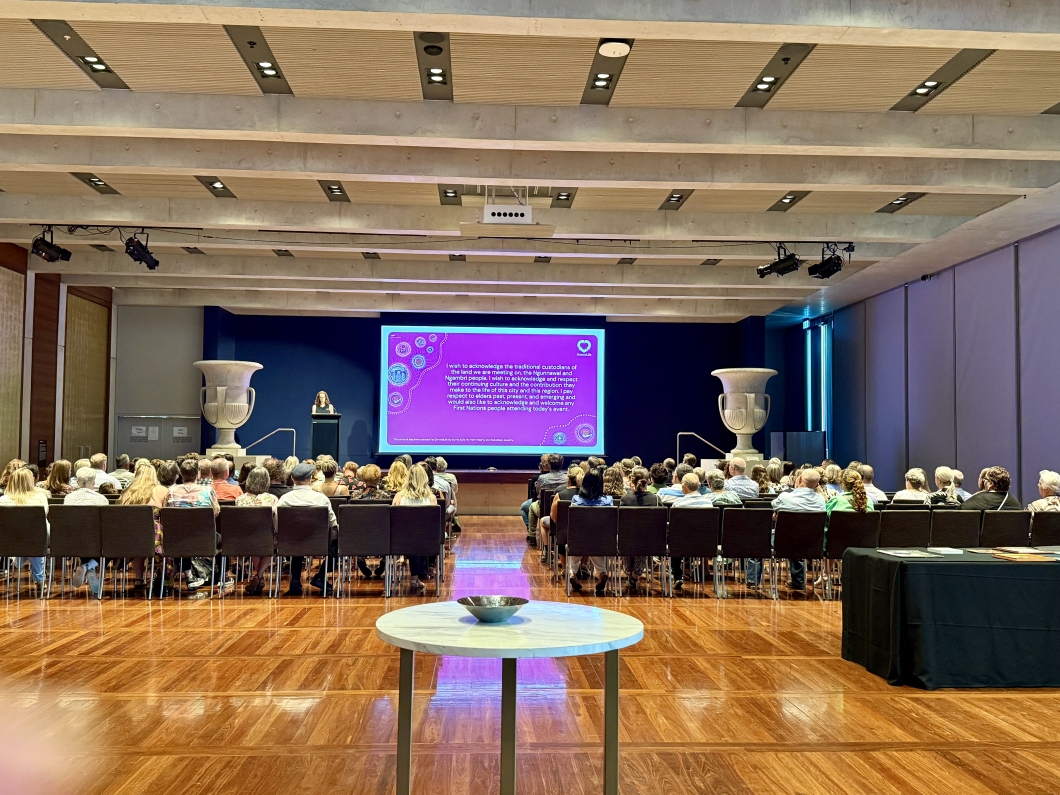 A large audience sits in rows facing a stage in a spacious hall with timber flooring. A speaker stands at a lectern beside a large screen displaying a bright magenta DonateLife-themed slide. The room features high ceilings with exposed beams, tall walls, and 2 large decorative urns positioned on either side of the stage. A round table is visible in the foreground, and a covered display table stands to the right.