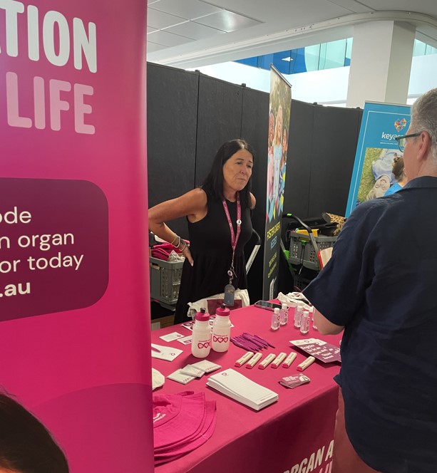 An information stall at an indoor expo featuring a magenta table with promotional materials, water bottles, and pamphlets about organ donation. A person stands behind the table speaking with another attendee, while large banners with partial text about organ donation and registration are displayed in the background.