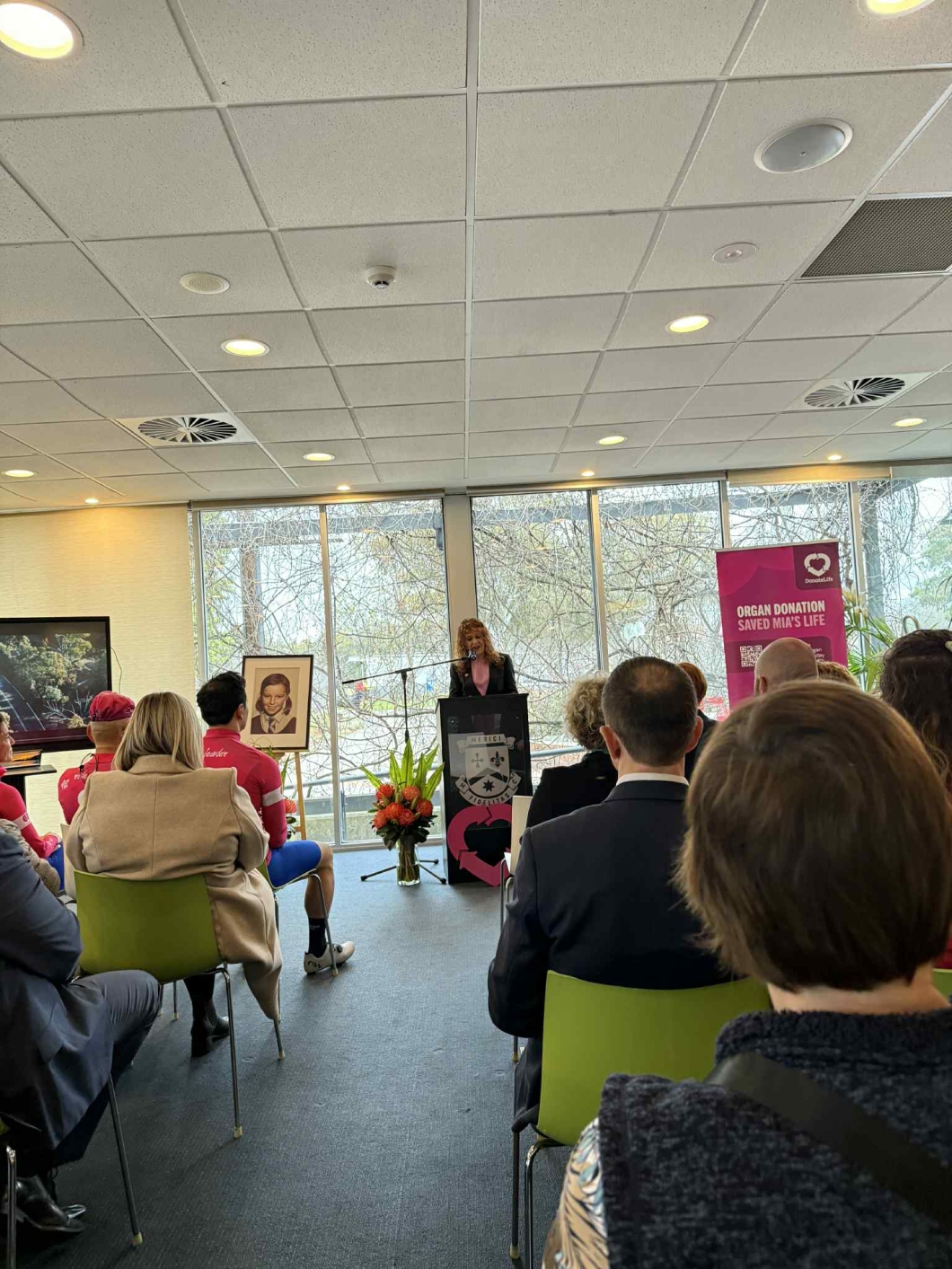 A woman is speaking at a podium in a room with large windows. The audience is seated facing her. A framed portrait on an easel is to the left of the speaker next to a vase of flowers, and a banner to the right reads ‘ORGAN DONATION SAVED MIA’S LIFE’. The room features modern decor with green chairs and ceiling lights.