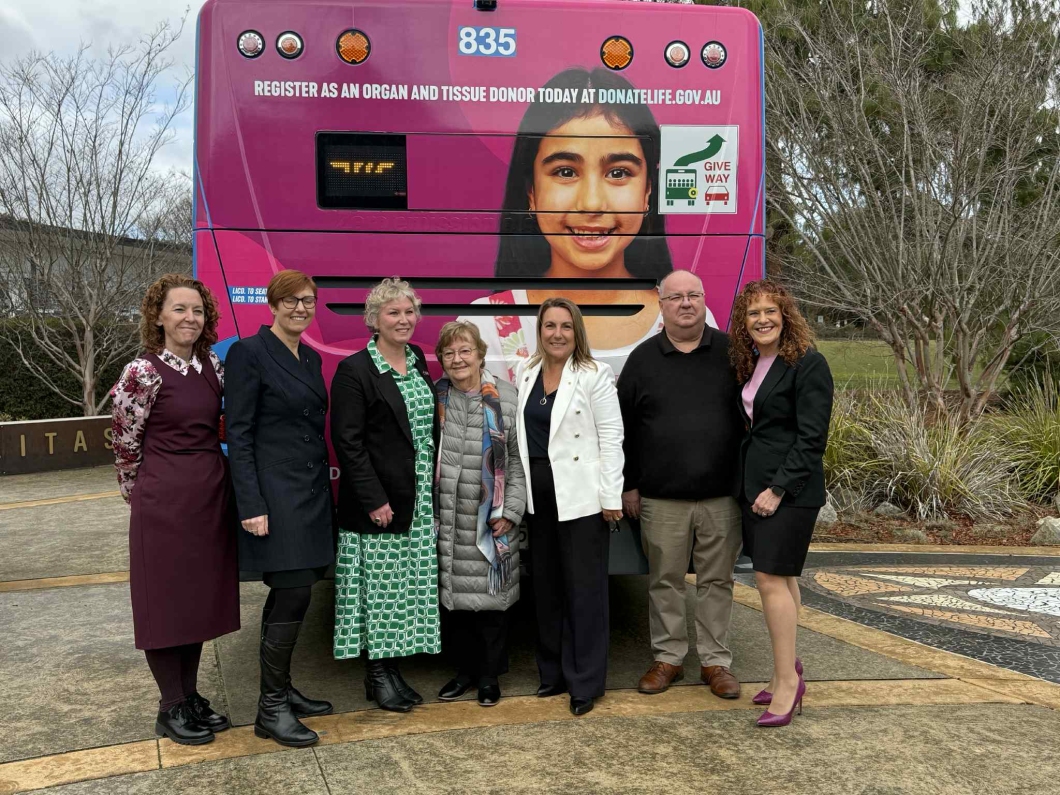 7 people stand in front of a magenta bus with the number 835. The bus displays a message promoting organ and tissue donation: ‘REGISTER AS AN ORGAN AND TISSUE DONOR TODAY AT DONATELIFE.GOV.AU’. Trees and a building are visible in the background.