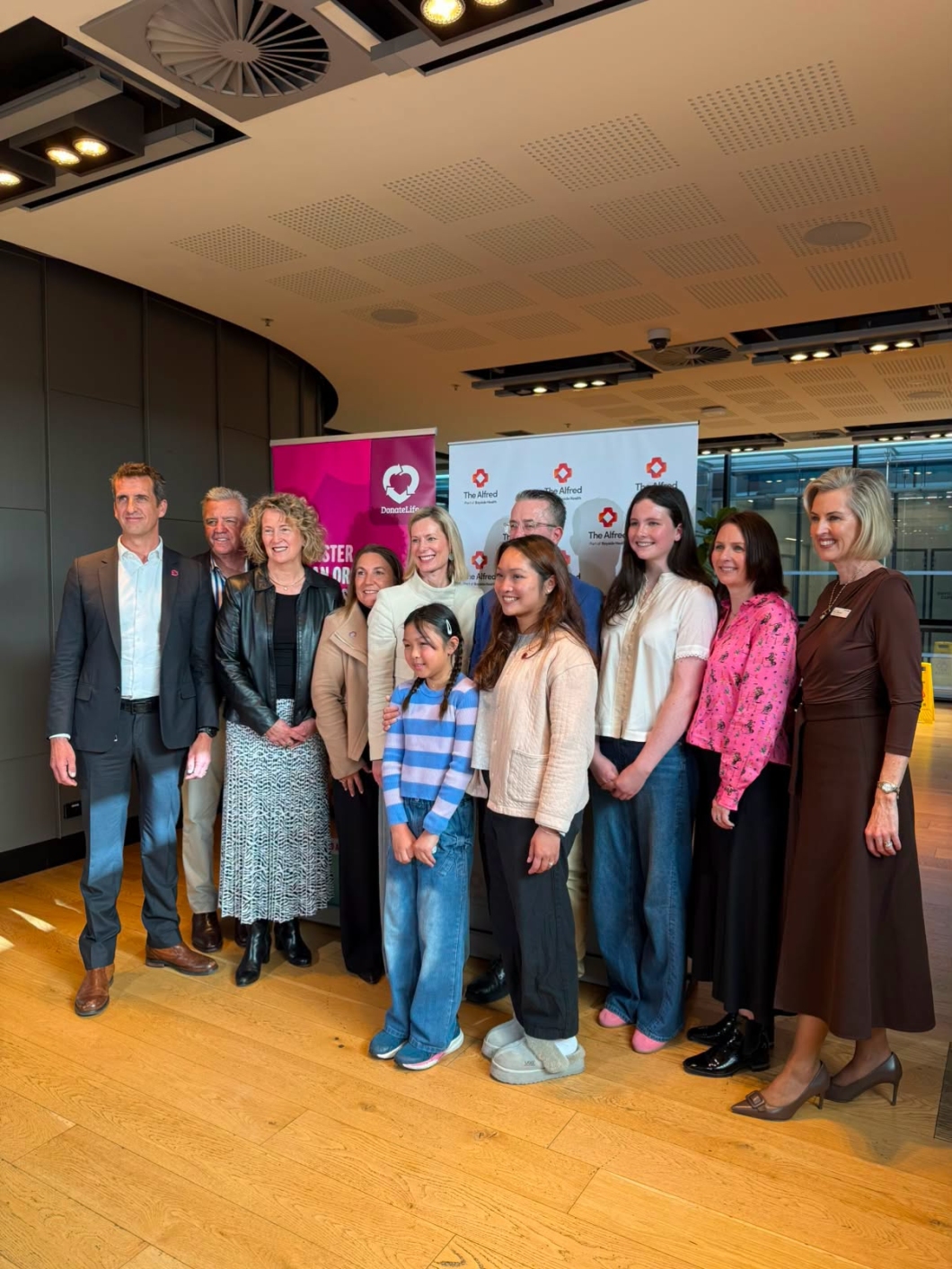 A group of 11 people stand together for a photo at an indoor event, in front of DonateLife and The Alfred banners. The group are arranged in 2 rows on a polished timber floor. The venue has high ceilings, modern lighting and large windows in the background. Visible signage includes the DonateLife logo and The Alfred logo.
