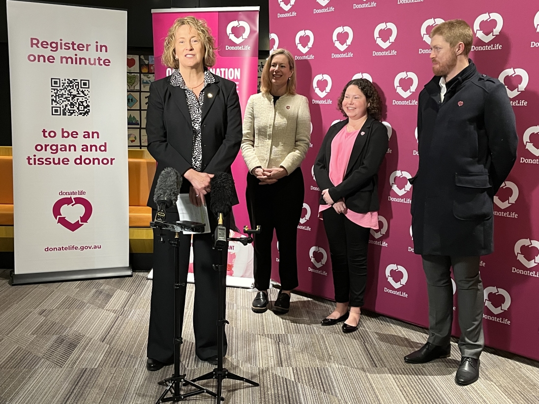 A group of 4 people stand in front of a backdrop with the DonateLife logo, which features a heart and the word 'DonateLife' repeated on a magenta background. One person is speaking at a podium with multiple microphones. To the left, there is a banner with the DonateLife logo at the top.