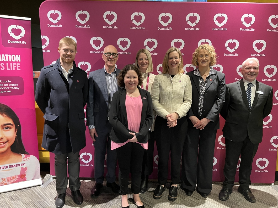 A group of 7 people standing in front of a magenta backdrop with the DonateLife logo, which features a heart and the words 'DonateLife.' To the left, there is a vertical banner with similar branding and text promoting organ donation. The individuals are dressed in business attire.     