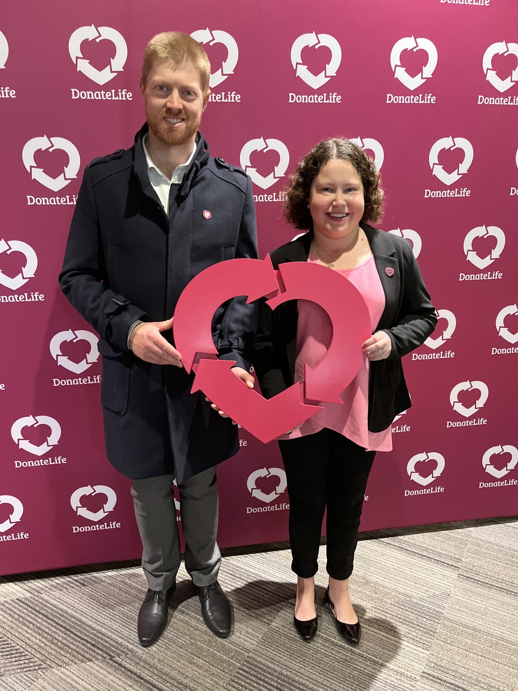 2 people standing in front of a backdrop with the DonateLife logo. The person on the left is wearing a dark coat and grey pants, while the person on the right is wearing a black jacket and pink shirt. They are holding a large magenta heart-shaped symbol together. 