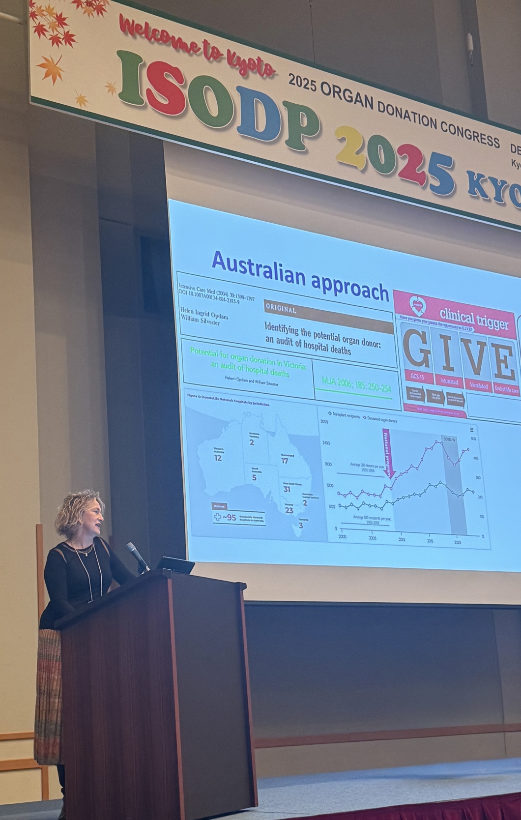 A presenter stands at a podium in a large conference hall, speaking beside a projected slide titled ‘Australian approach’. The slide includes maps of Australia, charts, and graphics related to organ donation. Above the stage hangs a large sign decorated with maple leaves that reads ‘Welcome to Kyoto – ISODP 2025 Organ Donation Congress Kyoto’.