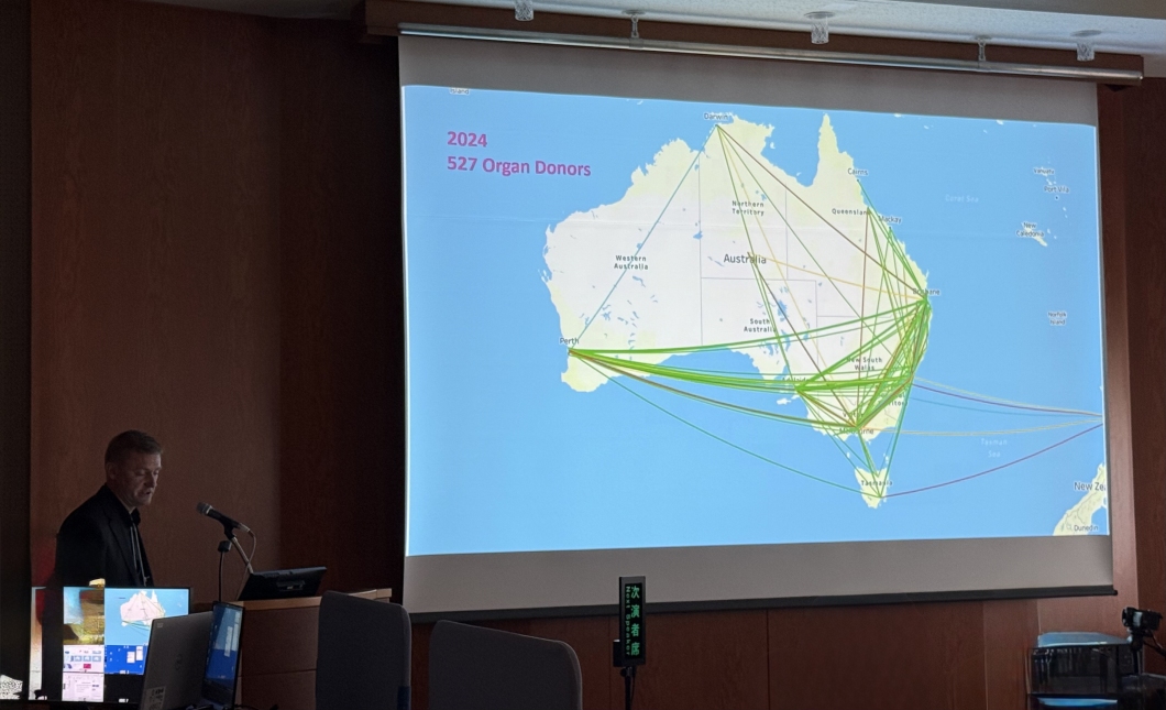 A person stands at a lectern in a conference room beside a large projection screen displaying a map of Australia. The map shows numerous coloured lines connecting different regions, illustrating movement or transfer pathways. Text on the screen reads ‘2024’ and ‘527 Organ Donors’. Several computer monitors and microphones are visible on the lectern in the foreground.