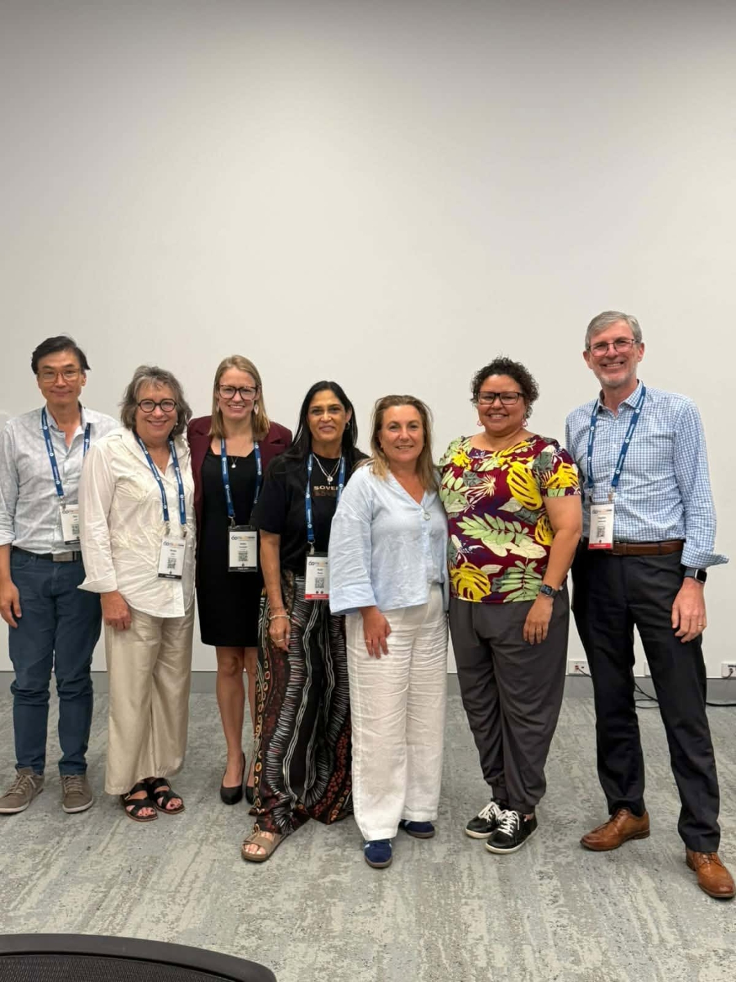 7 symposium delegates wearing event lanyards stand together for a group photo in a meeting room. The group is positioned against a plain white wall on a grey carpeted floor, wearing a mix of business and smart-casual attire.