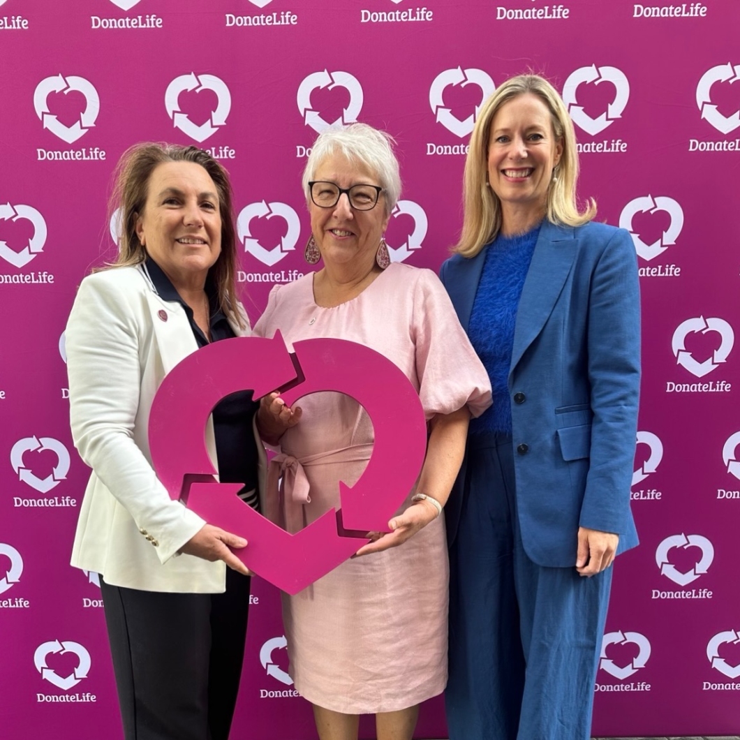 3 people stand in front of a magenta DonateLife media wall patterned with the DonateLife heart logo. They are holding a large pink DonateLife heart-shaped symbol together, positioned at the centre of the photo.