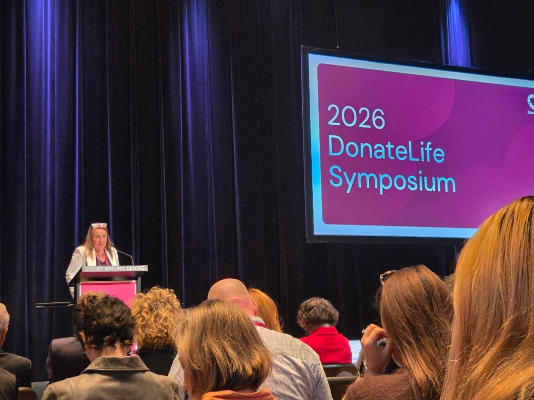 A speaker stands at a lectern addressing an audience at the 2026 DonateLife Symposium. Attendees are seated facing a stage with dark curtains, while a large screen displays the event title, ‘2026 DonateLife Symposium’. The presentation is taking place at the Sydney Masonic Centre.