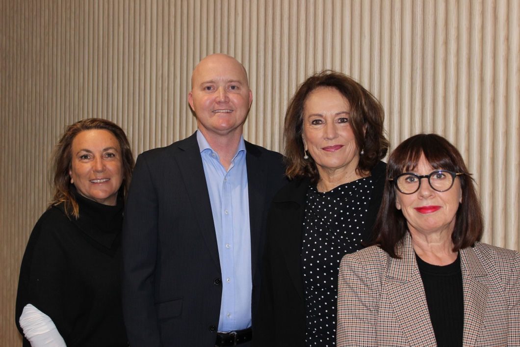 A group of 4 people standing close together indoors in front of a beige, vertically textured wall. One central person is wearing a dark suit jacket over a light blue shirt, while another next to them is wearing a dark top with a small white polka-dot pattern. The person on the left is dressed in a black top layered over a light-coloured long-sleeve shirt. The person on the right is wearing round glasses and a checkered jacket with a black top underneath. All 4 individuals are facing the camera.