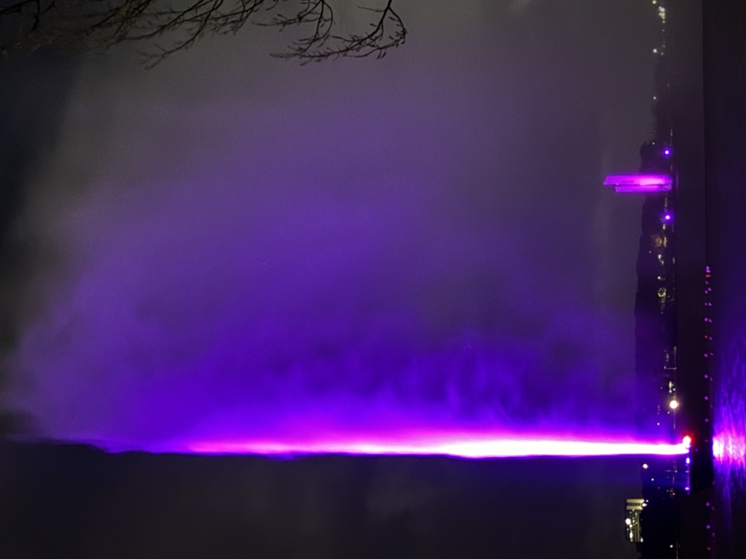 A vertical fountain illuminated with magenta light shines through mist against a dark background, with tree branches visible on the right side.
