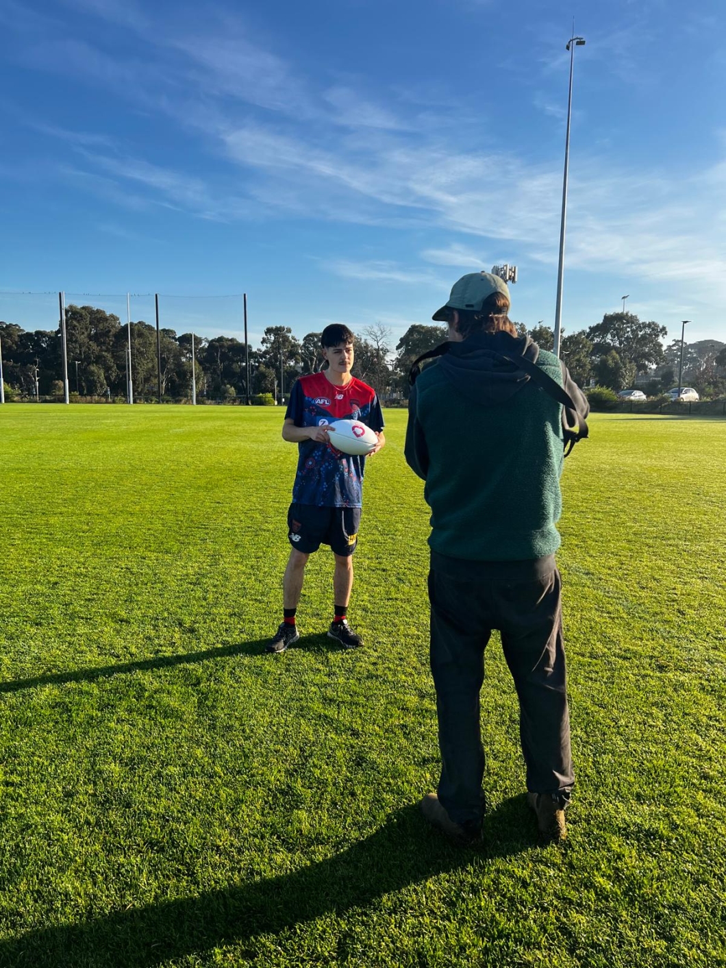 A person wearing a sports uniform stands on a grassy outdoor field holding an Australian rules football, facing a second person who is recording video. The second person is positioned with their back to the camera and holds a camera up to their face. The sports uniform includes a short-sleeved jersey with logos and dark shorts. The field is open and green, bordered by trees and tall light poles in the background under a clear blue sky. Long shadows stretch across the grass, indicating low-angle sunlight.