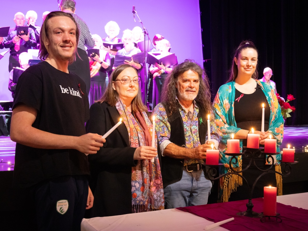 A group of people stand in front of a stage holding lit candles, with a choir performing in the background. They are behind a table with red candles on a candelabra. One person wears a black 'be kind' t-shirt, and another has long curly hair and colourful clothing.  