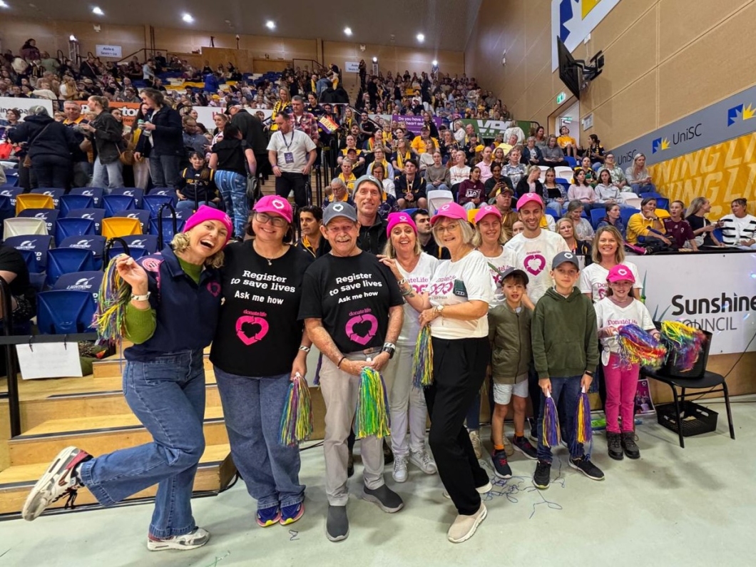 A group of people stand in front of a crowd at an indoor event or sports game, wearing matching 'Register to save lives Ask me how' shirts with a heart logo and 'DonateLife' text. Some hold colourful pom-poms. Spectators are seated in bleachers behind them, with visible sponsor banners including Sunshine Council and UniSC.