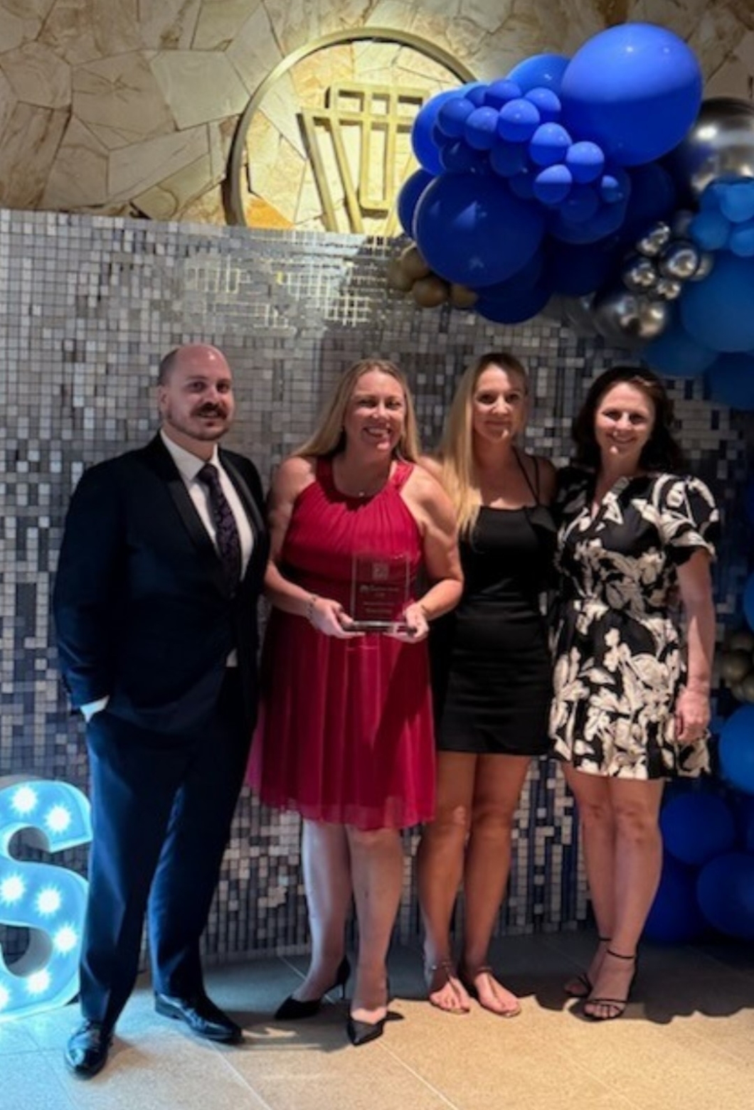 4 people stand in front of a decorative wall with blue and silver balloons. One person holds an award plaque. A large illuminated letter 'S' is on the floor to the left, and the background features a circular design with vertical bars and stone tiles.