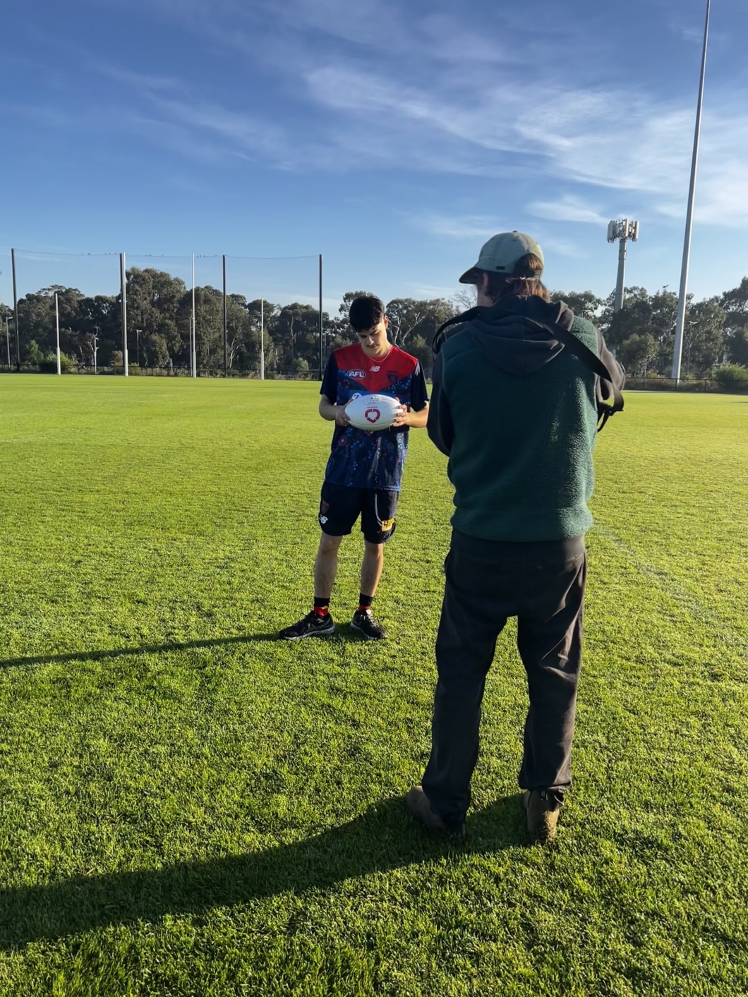 A camera operator films a person standing on a sports field and holding a white football featuring the DonateLife logo. The filming takes place on a sunny day, with goal posts, trees and a clear blue sky in the background.