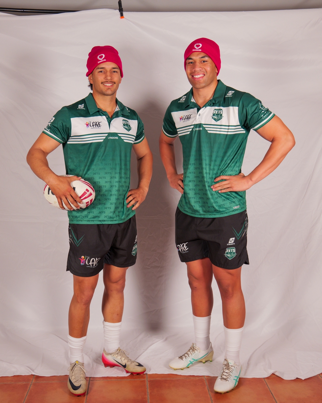 2 rugby league players stand in front of a white backdrop wearing green team uniforms and bright magenta DonateLife beanies. One player holds a rugby league ball while both pose with hands on hips.