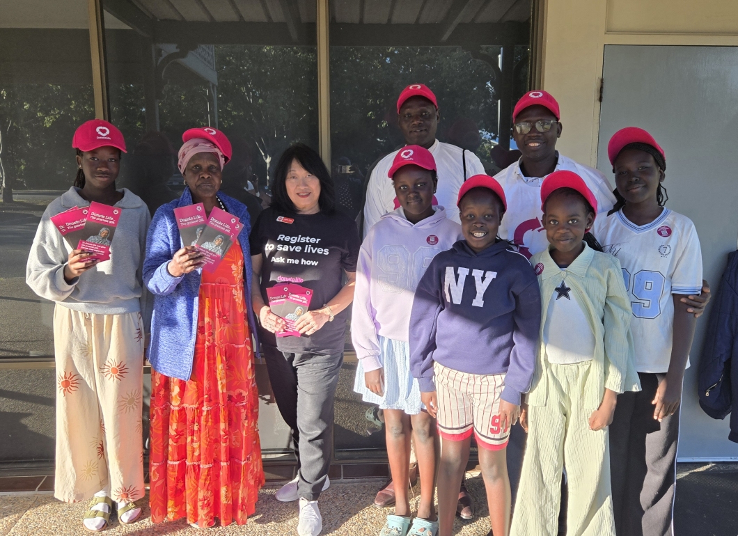 A group of community members wearing magenta DonateLife caps stands together outside a building, holding DonateLife information brochures. One person in the centre wears a black DonateLife shirt with the message ‘Register to save lives. Ask me how’. The group is gathered in bright sunlight in front of large windows as part of a DonateLife community awareness activity.