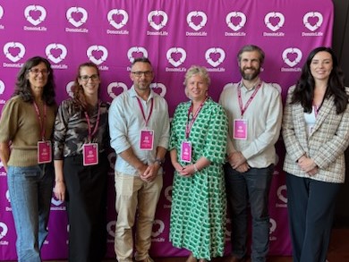A group of 6 individuals stand together in front of a pink backdrop with heart logos, wearing event badges.