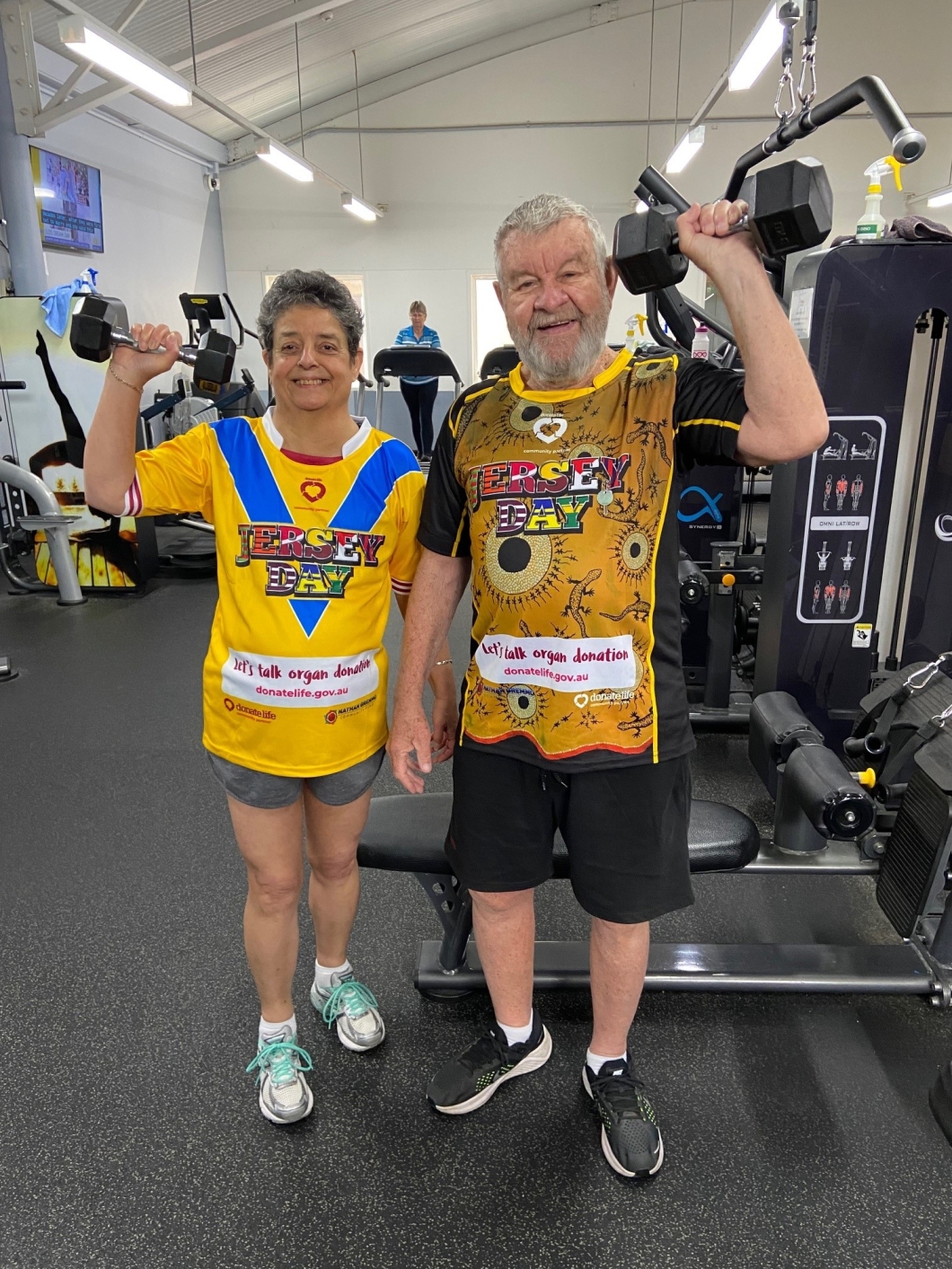 Two individuals in colourful ‘Jersey Day’ shirts hold dumbbells in a gym, promoting organ and tissue donation. Fitness equipment is in the background. shirts hold dumbbells in a gym, promoting organ donation awareness. Fitness equipment is in the background.